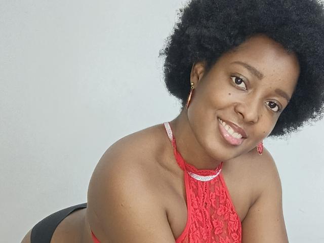 ChelseyeBonny with dark skin, afro, red lace halter top, showing her tits, smiling, red earrings, black panties, white background, looking at camera, pink lipstick, happy expression.