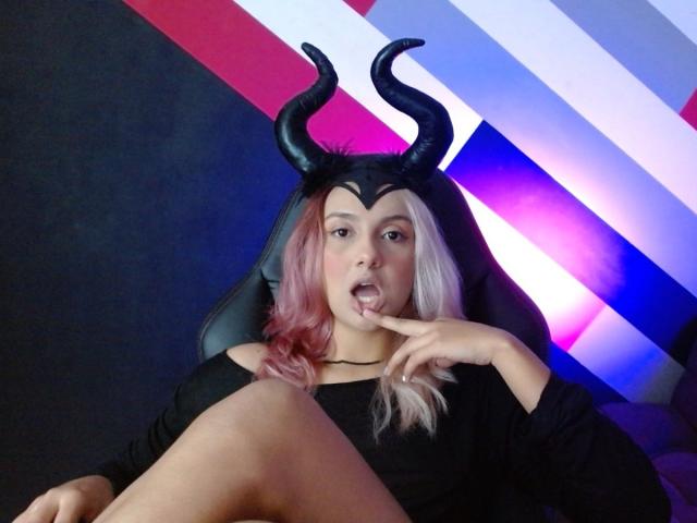 AlterEgoSecond with pink and white hair, black horns, black top, finger in mouth, sitting on chair, showing knee, colorful striped background, looking sexy, cosplay, slightly open mouth.