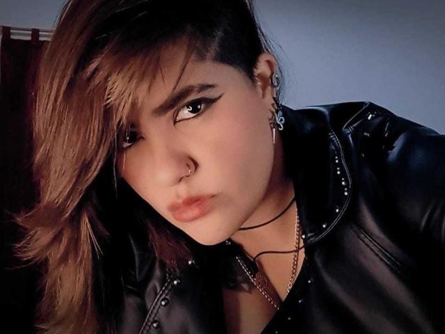 ZussanGravereign with brown hair, black leather jacket, nose ring, eyeliner, and serious expression, wearing skull earrings and choker, looking directly at camera.