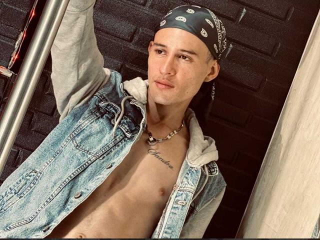 PosionAngel with a skinny, shirtless build, wearing a blue denim vest, black bandana, necklace, and grey hoodie, standing by a pole, looking at the camera.