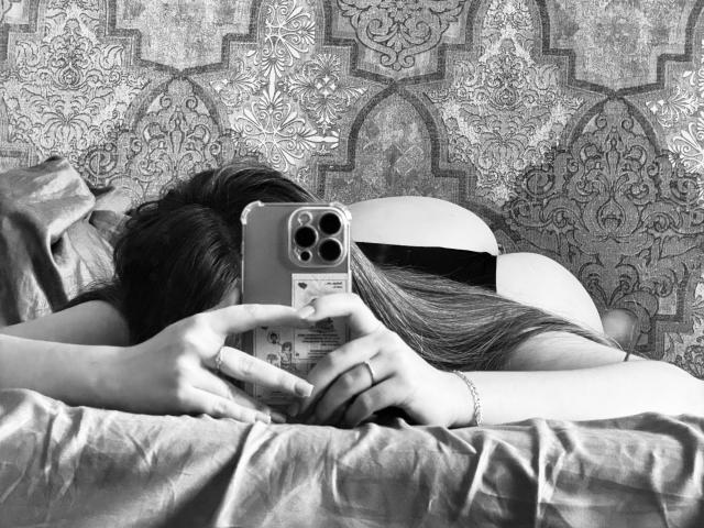 ElisaHotlis lying on bed, face down, taking a selfie with a phone. Her long hair covers her ass. She's wearing a ring and a bracelet. Black-and-white photo.