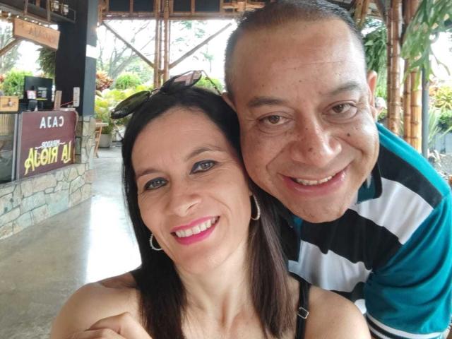 GabrielAndAbygail with short-cropped bald man, smiling, arms around each other's shoulders. She has pink lipstick, black hair, hoop earrings, he's in green-striped shirt. Both happy, brown eyes.