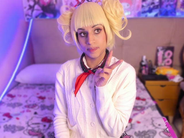 EimyFlorian mit blonden Zöpfen, die einen weißen Pullover und ein rotes Halstuch trägt, zeigt ein Friedenszeichen und präsentiert ihr süßes Gesicht und ihre kleinen Brüste in einem gemütlichen Schlafzimmer..