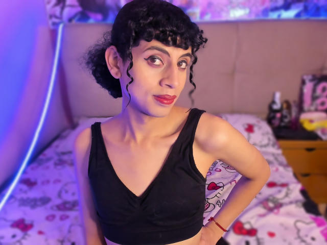 EimyFlorian with curly black hair, red lipstick, and black tank top, showing small tits and a slim, pale-skinned body, hands on hips, in a bedroom.