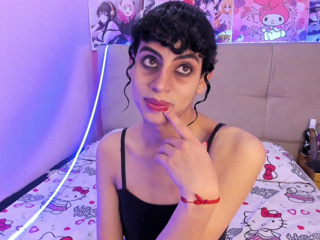 EimyFlorian with curly black hair, dark makeup, and red lipstick, finger in mouth, black tank top, red bracelet, sitting on Hello Kitty bed, looking up with big eyes.