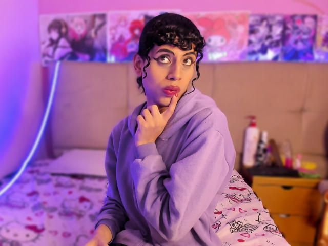 EimyFlorian with curly black hair, purple hoodie, finger near pink lips, wide-eyed, on a bed with Hello Kitty pattern, purple and blue lights in the background.