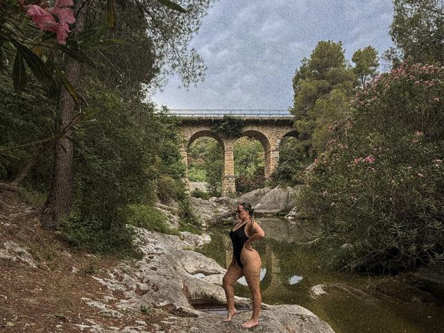 MonaPlus stands on a rocky riverbank in a black one-piece, showing her ass and bare pussy, with a stone arch bridge in the background and greenery around.