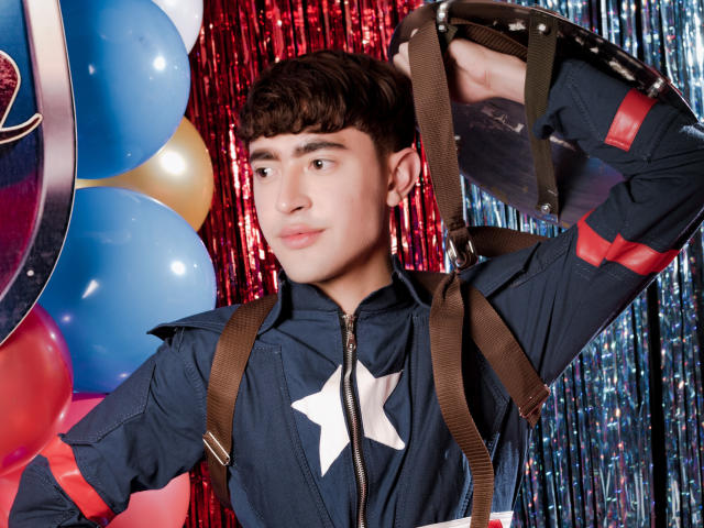 JustinStone with short brown hair, wearing a navy zip-up with a white star, brown suspenders, and a drum over his shoulder, looking slightly to the side with a calm expression.
