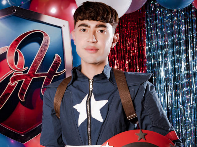 JustinStone with short brown hair, wearing a blue zip-up jacket with a white star, brown suspenders, holding a red star, surrounded by balloons and shiny red and blue streamers.