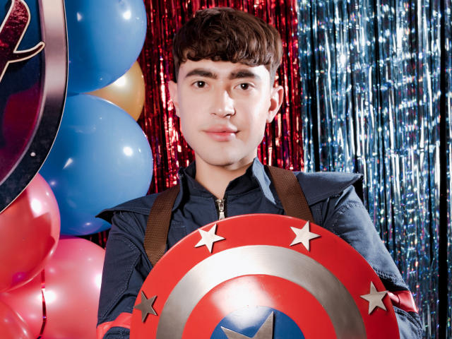 JustinStone, a young dude with brown hair and a nose ring, holds a Captain America shield, wearing a blue jacket, surrounded by balloons and shiny silver curtains.