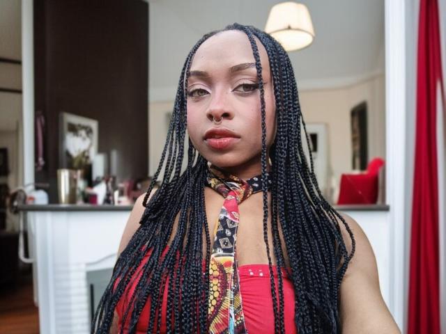 QueencyBraxton with long black braids, wearing a red top and colorful scarf, has full lips, light brown skin, and a serious expression. Her tits are medium-sized and she's looking straight at the camera.