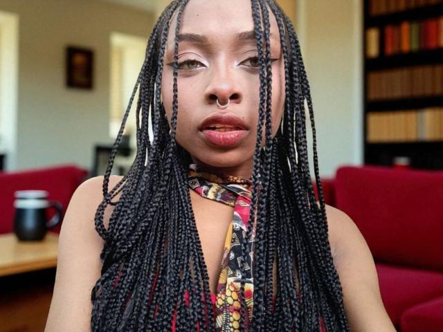 QueencyBraxton with long black braids, nose ring, and a floral-patterned top, looking directly at the camera with slightly parted pinkish lips, in a red sofa-filled room.