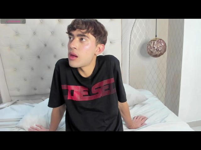 JustinStone with curly brown hair, wearing a black t-shirt with red stripes, sitting on a bed, looking to the side with a surprised expression, hands on white pillows.