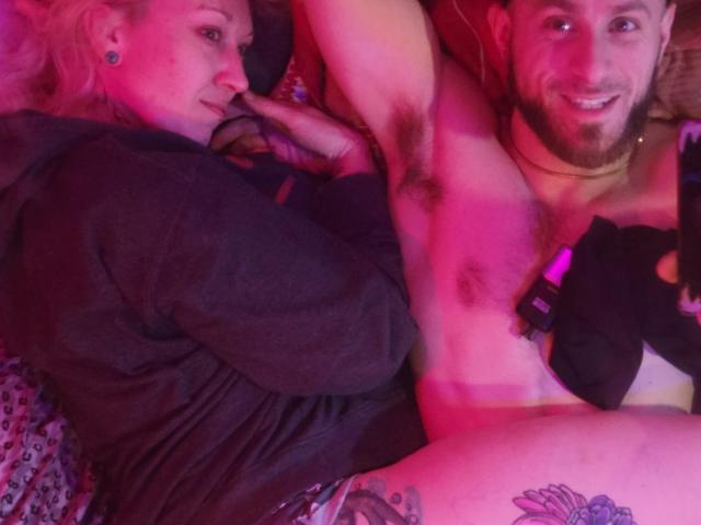 CloudyCouple's topless dude with a beard and chest hair, holding a phone, lying next to CloudyCouple's pink-haired woman with tattoos, in pink bedroom light.