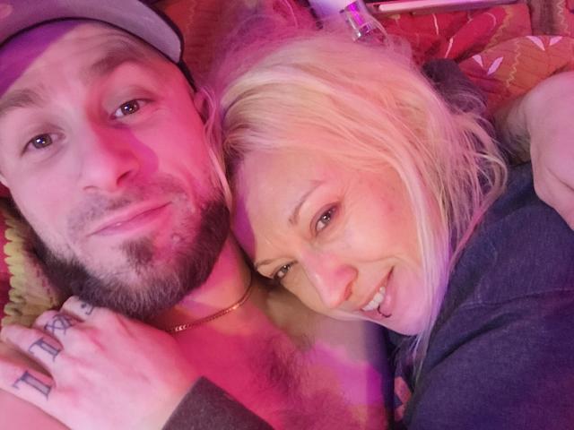 CloudyCouple has a cute, smiling blonde woman with tits and a bearded man with a cock ring on his finger, lying down under pink and purple lighting.