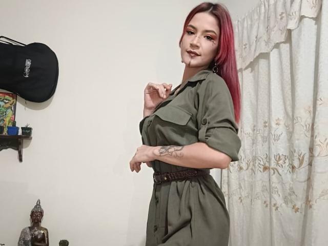 CherryDickens with red hair, green dress, and brown belt, holding her shirt, showing tattoo on her right arm, standing in a room, looking over her shoulder.
