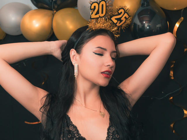 LizzeJoy with black hair, wearing a gold "25" crown, black lace top, red lipstick, and gold earrings, has arms raised, showing her tits, with eyes closed, looking sexy.