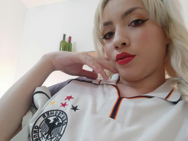 HaileyDavies with blonde hair, red lipstick, and a white soccer shirt with black, red, and yellow stars, resting her chin on her hand, looking at the camera.