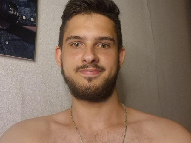 Lumut has a shirtless, hairy chest, short brown hair, and a beard, wearing a silver necklace, with a slight smile. His brown eyes are centered in the image.