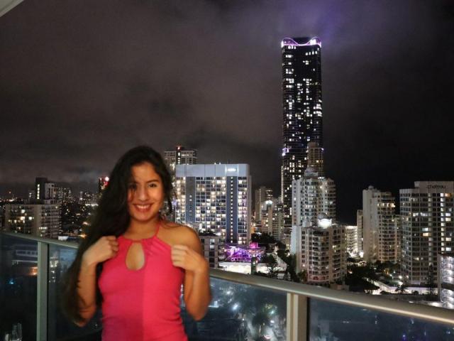 Aurora Monroe mit langen, dunklen Haaren, lächelnd, trägt ein pinkes Top mit Trägern und einem Ausschnitt, steht auf einem Balkon in der Nacht, Stadtansicht mit hohen Gebäuden und violetten Lichtern..