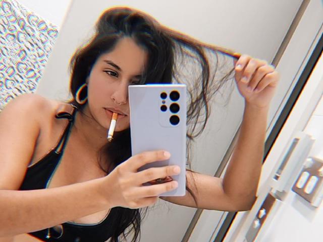 AuroraMonroe with long dark hair, holding a phone with a cigarette in her mouth, wearing a black tank top, large hoop earrings, and a nose ring, looking at the camera.