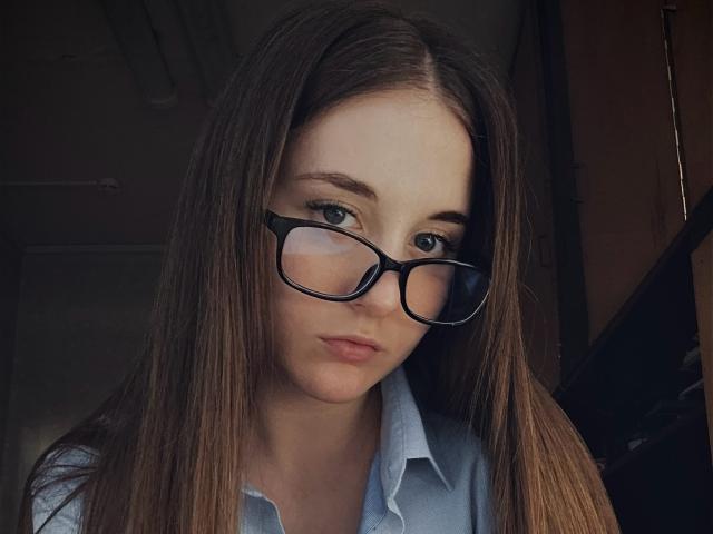 ElisaHotlis has long brown hair, black glasses, and a serious expression, wearing a blue shirt, with pale skin and subtle makeup. Her tits are small and her pussy is unseen.