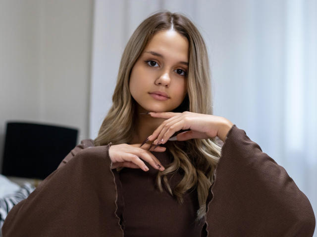 AgnesBelle with long, wavy brown hair, brown eyes, and a cute face, wearing a brown top with wide sleeves, touching her chin with her fingers, looking at camera.