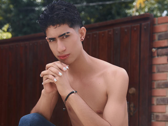 NovaWheeler, shirtless, with curly black hair, brown skin, and a slight tan, has white nail art, a black bracelet, and a stud earring, sitting with hands near his face.