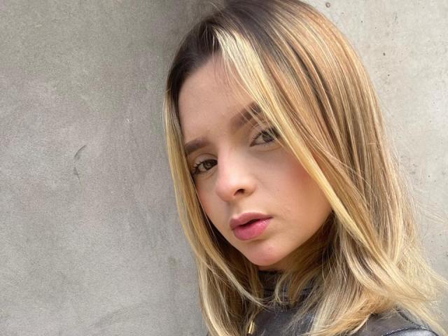 PauliAdams has blonde, straight hair, fair skin, and pink lips. She's looking to the side with a neutral expression, wearing a gray top. Her tits are not visible.