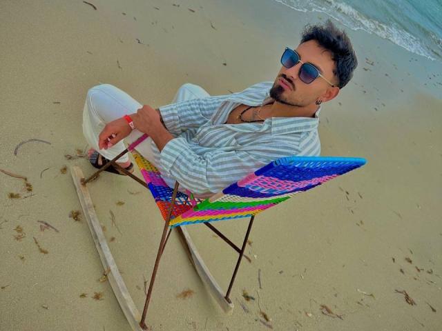 IvannaAndFriend, wearing a striped shirt and white pants, sits in a colorful folding chair on a sandy beach, sporting aviator sunglasses and a beard, with a relaxed expression.