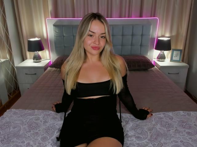 Eva Adderiy, con cabello rubio, una blusa negra con un hombro descubierto y una falda negra, sentada en una cama, mostrando sus pequeños senos, su cara linda y sus labios rosados, con un peinado morado neón..