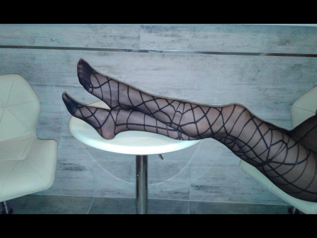 Sophia01-hot's legs in black, criss-cross patterned pantyhose, crossed at the ankles, resting on a white, circular table with white chairs on either side.