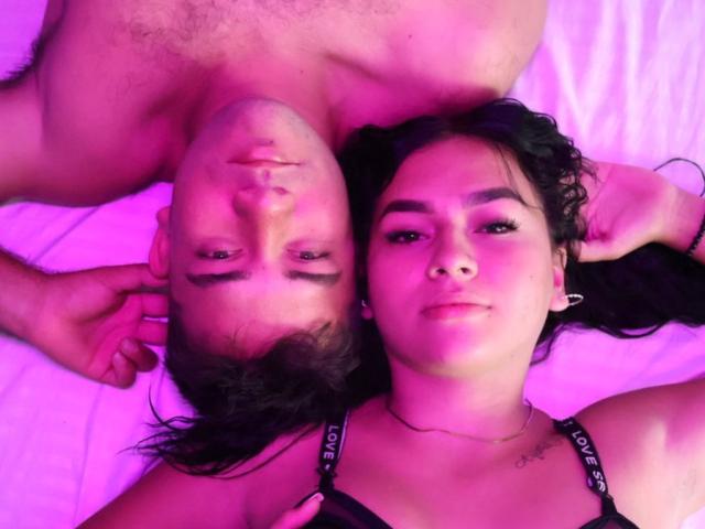 CharlotteAndTheo's face is next to Theo's, both lying on a bed with pink lighting. Theo's shirtless, CharlotteAndTheo has dark hair and a black top.