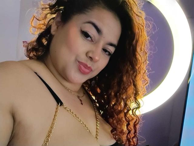 Lalitzfet, con i capelli castano-arancio ricci, indossa un reggiseno a bretelle nero, una catena dorata e rossetto rosa, sorridendo dolcemente alla fotocamera. Il suo seno è visibile, con un'espressione sottile e sicura..