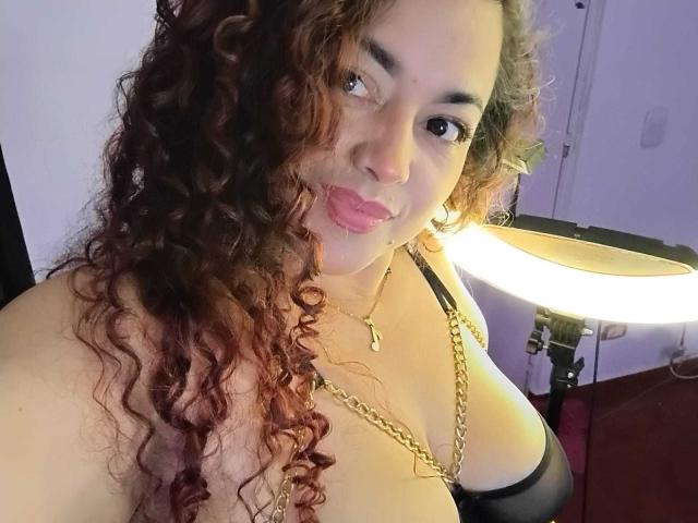 Lalitzfet con capelli castano ricci, labbra rosa, collana d'oro, reggiseno nero, seno abbondante, che guarda la fotocamera, luce ad anello a destra, luce soffusa, incarnato caldo, espressione leggermente giocosa..