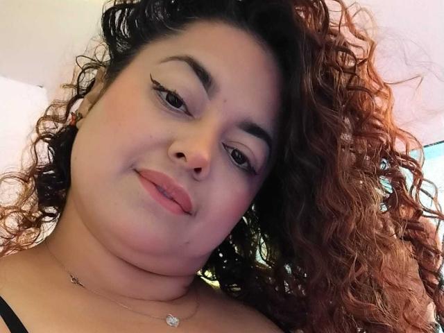Lalitzfet a les cheveux bruns bouclés, des sourcils foncés, et elle sourit avec du rouge à lèvres rose. Elle porte un haut noir avec un petit collier. Son nez est percé..