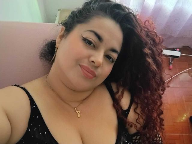 Lalitzfet with curly red hair, wearing a black, sparkly, low-cut top, showing her big, round tits and a gold necklace with a "B" pendant, smiling slightly.