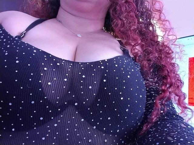 Lalitzfet's big boobs in a black, polka-dotted, ribbed top with spaghetti straps, showing a bit of cleavage. She has curly red hair and a small necklace.
