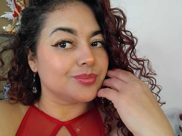 Lalitzfet with curly red hair, wearing red lipstick and a red halter top, touching her cheek with a slight smile, brown eyes, and small earrings.