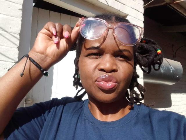 RounderAss with dark skin, puffy pink lips, and small nose, wearing pink sunglasses, holding them up, has black braided hair with colorful ends, and a black shirt.
