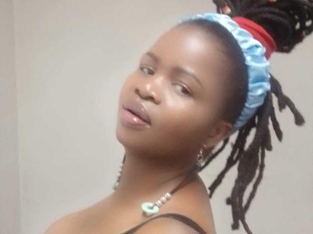 RounderAss with shiny brown skin, shiny lips, blue satin headwrap, black dreadlocks, necklace, and earrings, smiling slightly, showing off her small, cute tits.