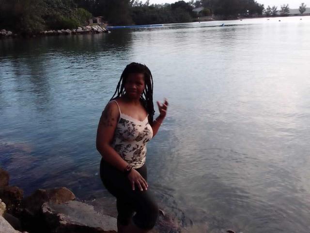RounderAss with dark skin and braided hair stands in shallow water, wearing a white floral tank top and black pants, touching her hair with a slight smile.