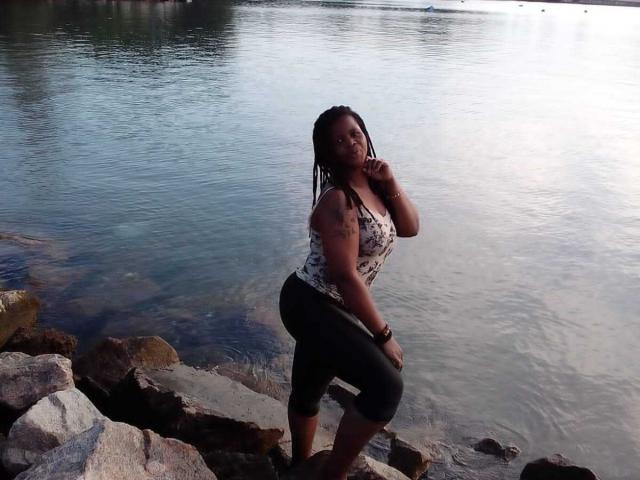 RounderAss with dark skin and braided hair, wearing a patterned top and black capris, standing on rocks by a calm water body, touching her chin.
