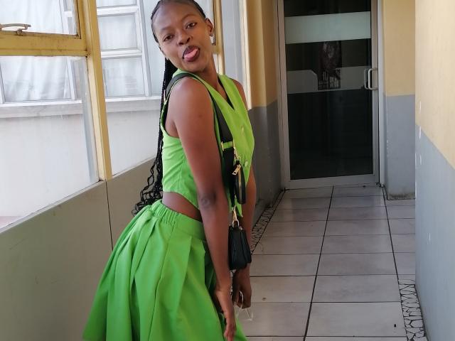Liisay with dark skin, long braided hair, green dress with cutout sides, standing in hallway, black purse, tongue out, looking back, cute, attractive, small breasts.