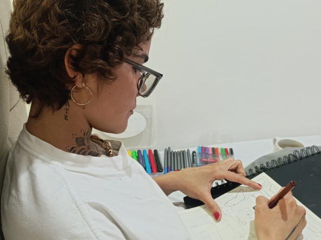 AnaRoa, con cabello castaño rizado, gafas, aros grandes, camisa blanca, tatuaje en el cuello, dibujando en un papel con un lápiz, bolígrafos de colores en un portalápices, mano derecha apuntando, mano izquierda sosteniendo el lápiz..