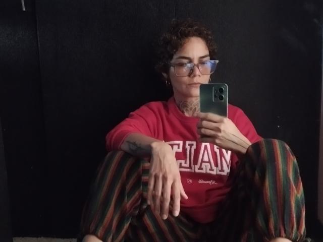 AnaRoa, con i capelli castano ricci, occhiali, una maglietta rossa, pantaloni a righe, un tatuaggio sull'avambraccio destro, che si fa un selfie con un telefono verde, seduta con le gambe sollevate, con un leggero sorriso, su uno sfondo scuro..