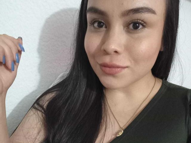 MartinaMarX with long black hair, light brown skin, and blue nails, wearing a green shirt and gold necklace, smiling softly at the camera.
