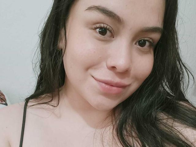 MartinaMarX has pale skin, brown eyes, and black wavy hair, smiling with a slight nose bump and natural makeup, wearing a black spaghetti strap top.