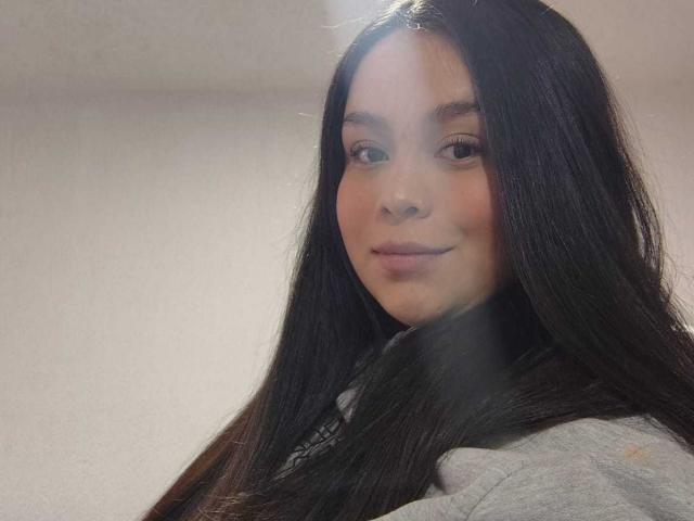MartinaMarX has long, straight black hair, a cute face with subtle makeup, and a small, confident smile. She's wearing a gray hoodie, showing a relaxed, pretty look.