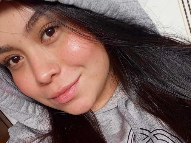 MartinaMarX hat dunkle braune Augen, hellrosa Lippen und langes schwarzes Haar. Sie trägt einen grauen Kapuzenpullover, hat ein leichtes Lächeln und ein paar kleine Sommersprossen auf der Wange..